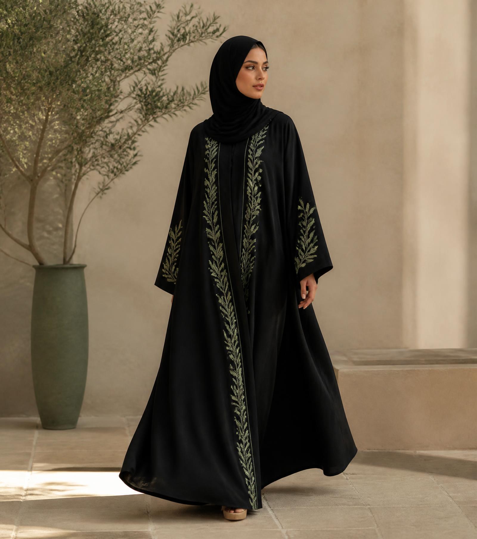 Model wearing an embroidered black abaya beside an olive tree